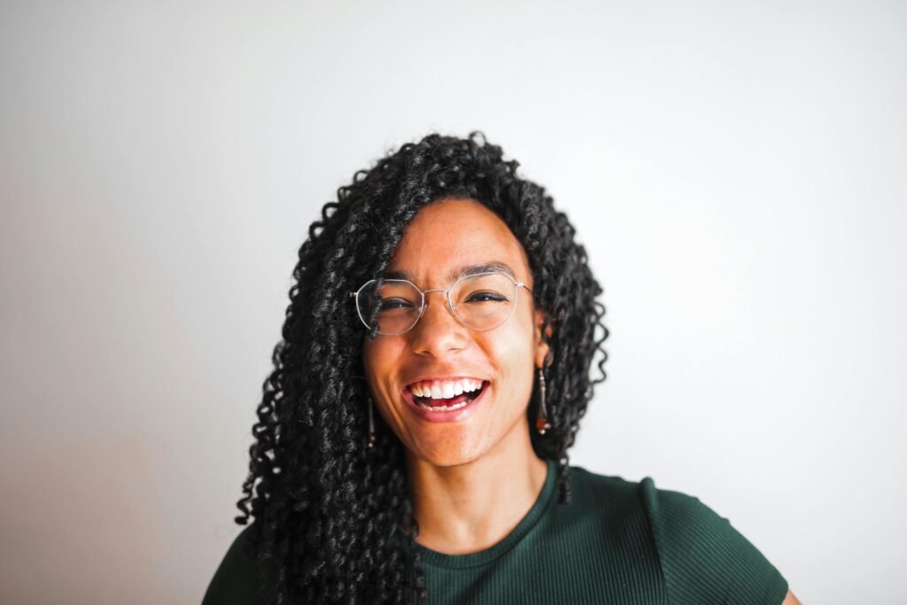 A joyful portrait of a young woman with glasses and curly hair smiling against a simple white background.